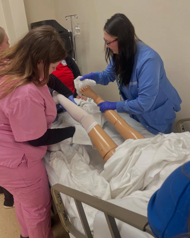 Compass Career College Allied Health students practice bandaging on a training mannequin's legs in a clinical setting.