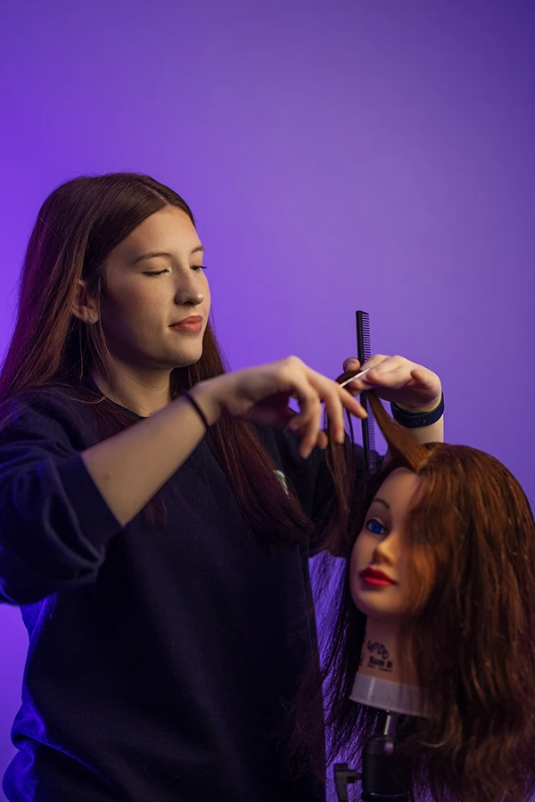 Young woman practicing hair cutting on a mannequin head with long brown hair and a purple background as a Cosmetology student at Compass Career College.