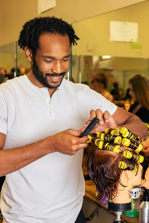 Man styling a mannequin head with wet hair and yellow curlers in a salon setting as a Compass Career College Cosmetology student.