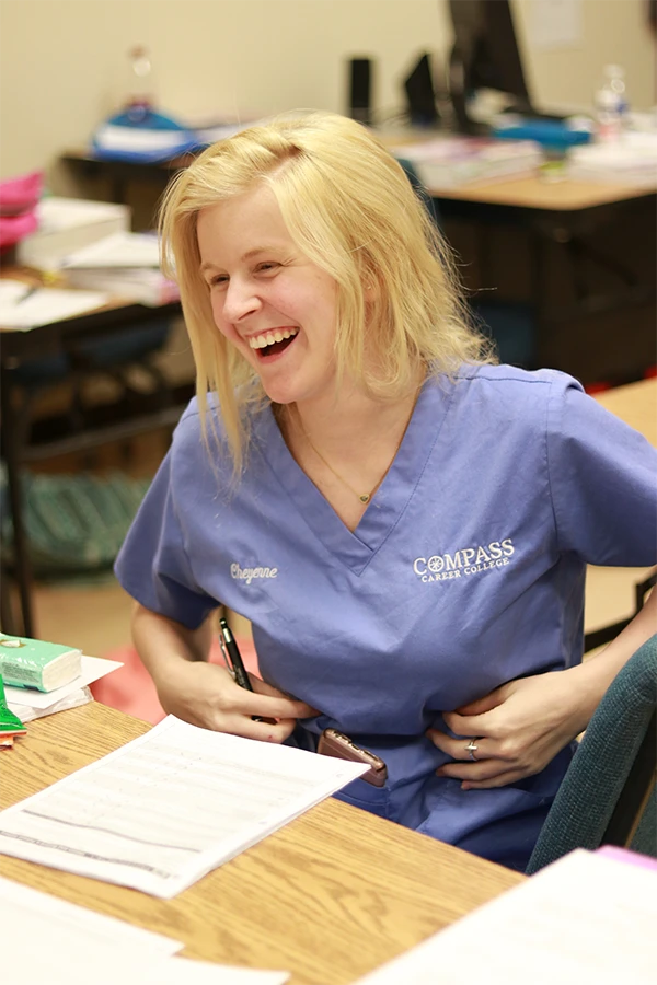 Smiling woman in blue scrubs sitting at a desk with papers and pen at Compass Career College.