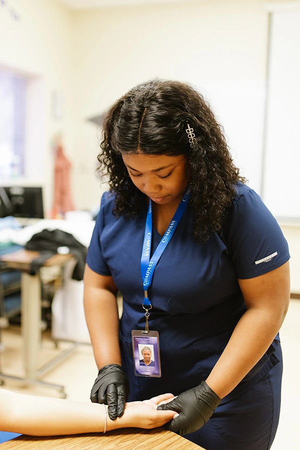 A Compass Career College LPN student in navy scrubs and gloves checks a patient's wrist pulse in a clinical setting.