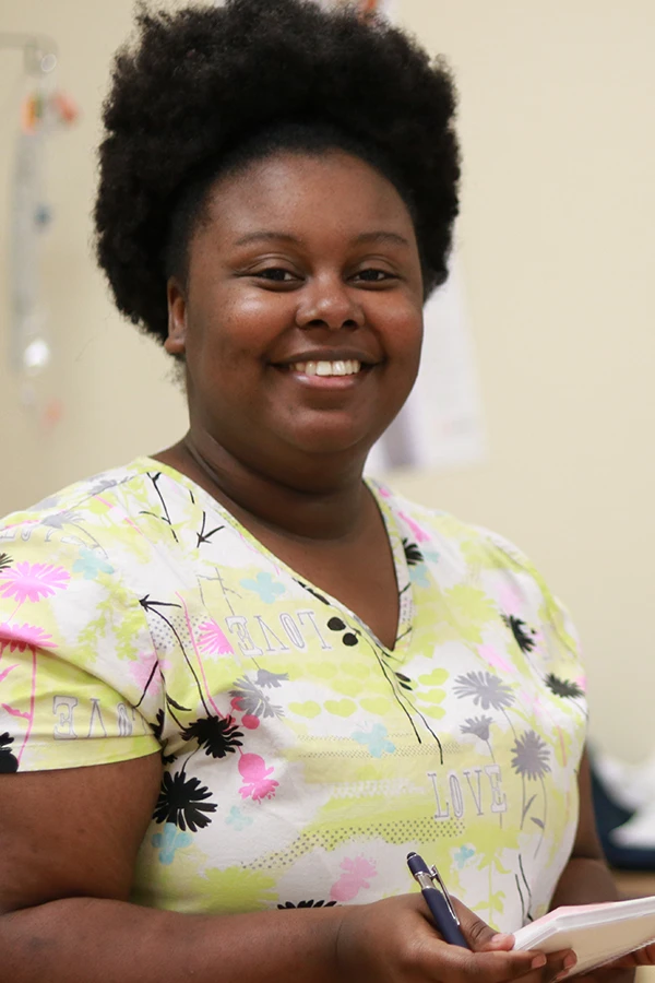 A smiling woman with an afro wearing floral-patterned scrubs, holding a pen and notepad as an Allied Health student at Compass Career College.