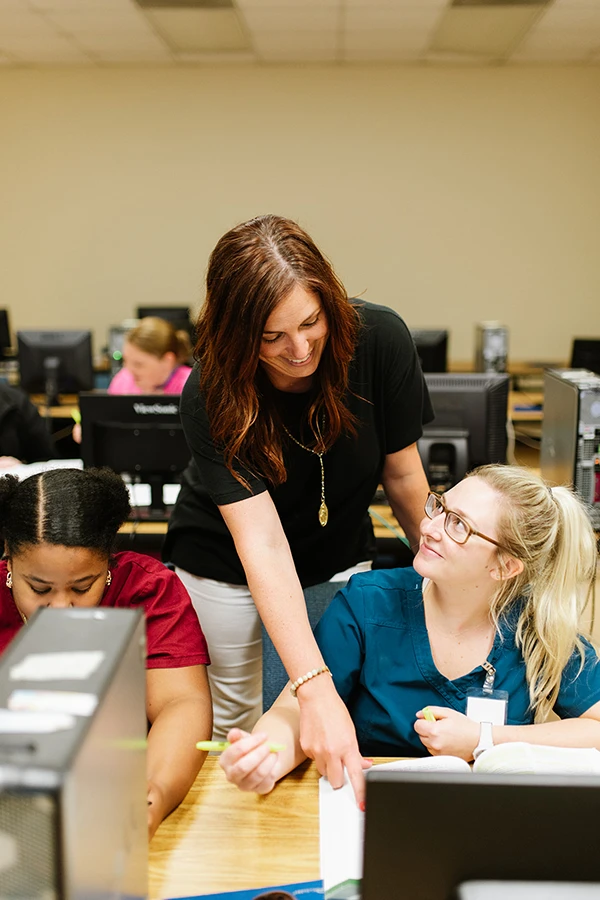Compass Career College instructor helps a student in blue scrubs at a computer in a classroom with other students.
