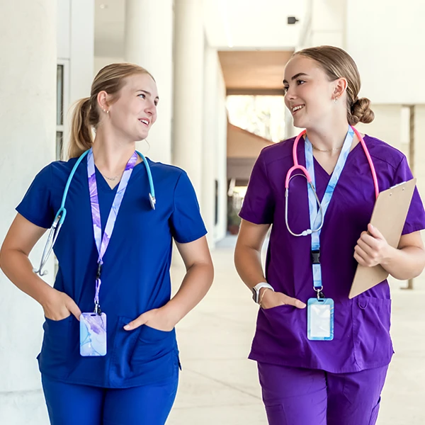 Two nurses in blue and purple scrubs walking and talking with stethoscopes and ID badges.
