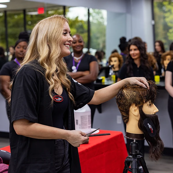 Compass Career College graduate hairstylist smiling while styling hair on a mannequin head in a salon classroom.