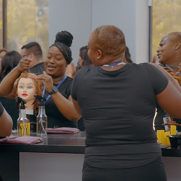 Two Compass Career College students styling mannequin heads in a hair salon with spray bottles on the counter.