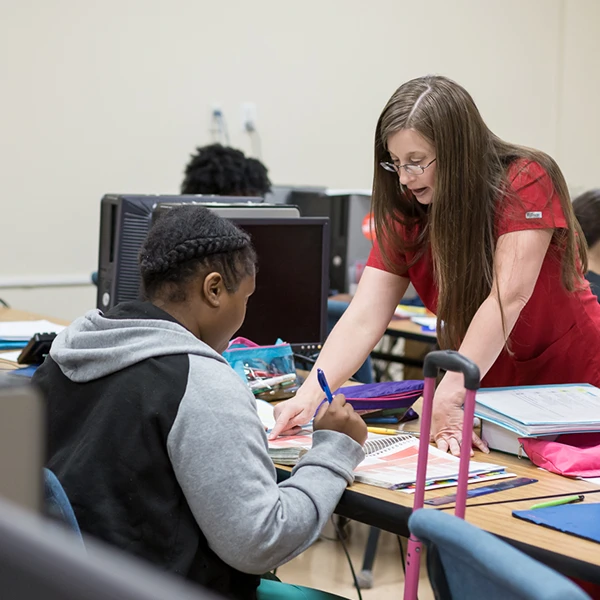 Compass Career College instructor in red helping student with notebook work at a classroom desk with computers around.