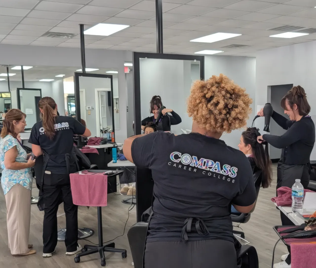 Three hairstylists working on clients' hair in a salon with Compass Career College logos on their shirts.