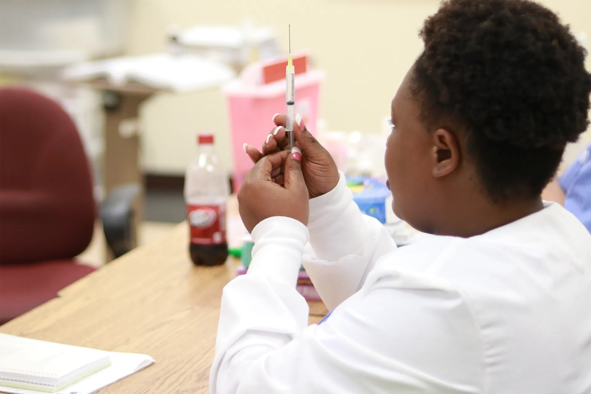 Compass Career College Allied Health student healthcare worker in white coat holding a syringe filled with liquid in an office setting.
