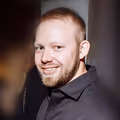 Vadim Tsyplakov s krátkými vlasy a vousy v černé košili na tmavém pozadí.