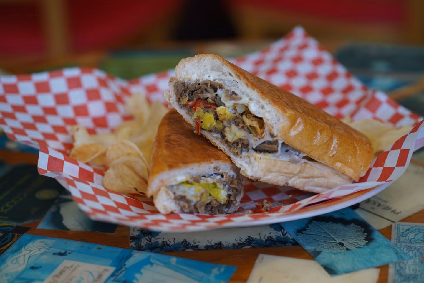 Philly cheesesteak sandwich with meat, cheese, and peppers served with potato chips on checkered paper.