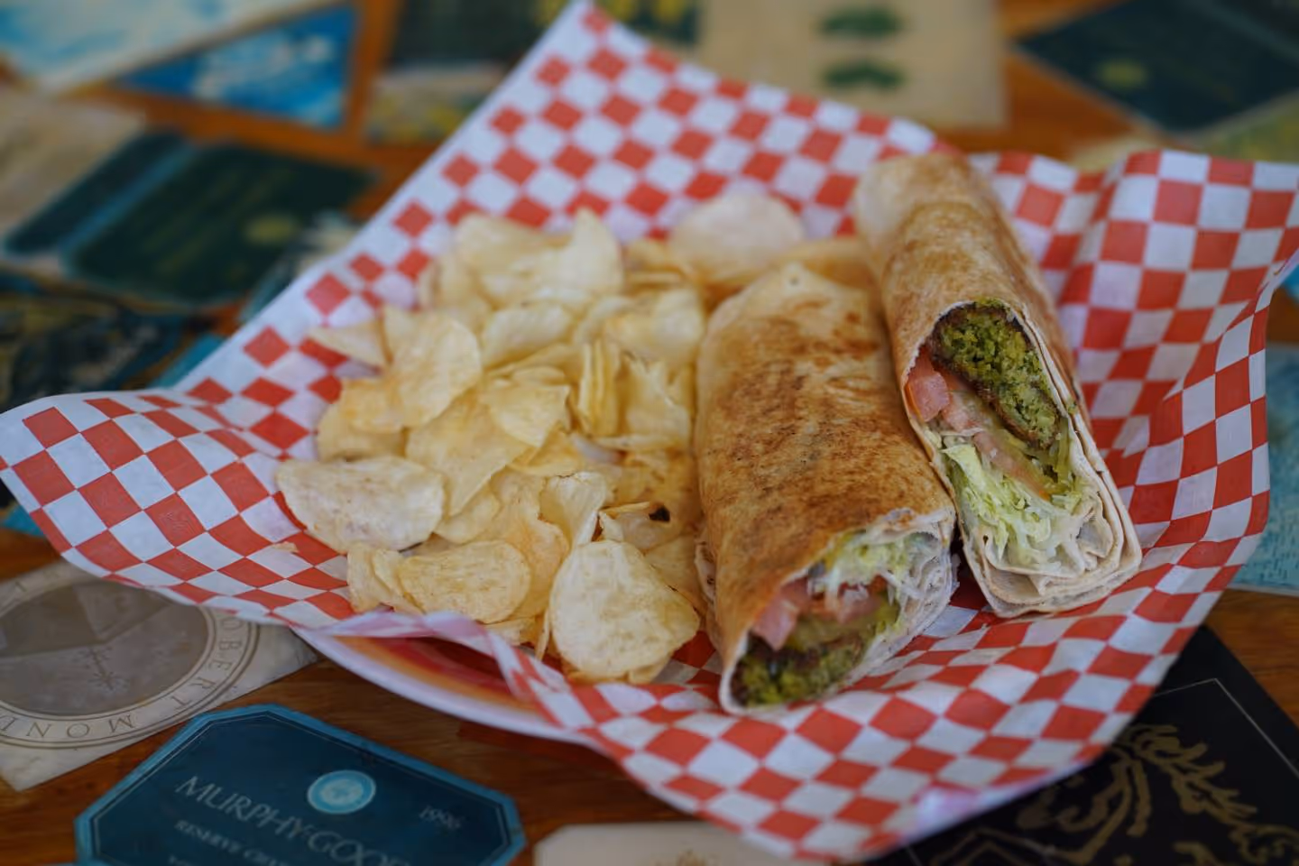 Plate with falafel wrap halves and a side of potato chips on red and white checkered paper.