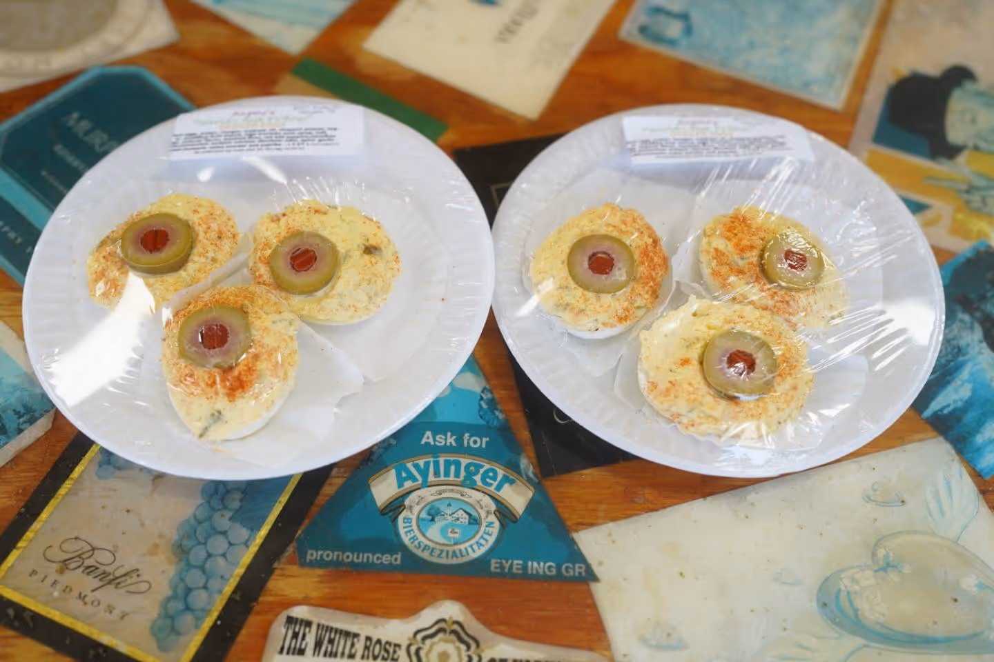 Two plates with three deviled eggs each topped with green olive slices wrapped in plastic.