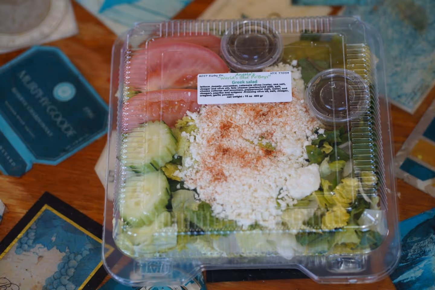Packaged Greek salad with sliced tomatoes, cucumbers, feta cheese, and two small dressing containers.