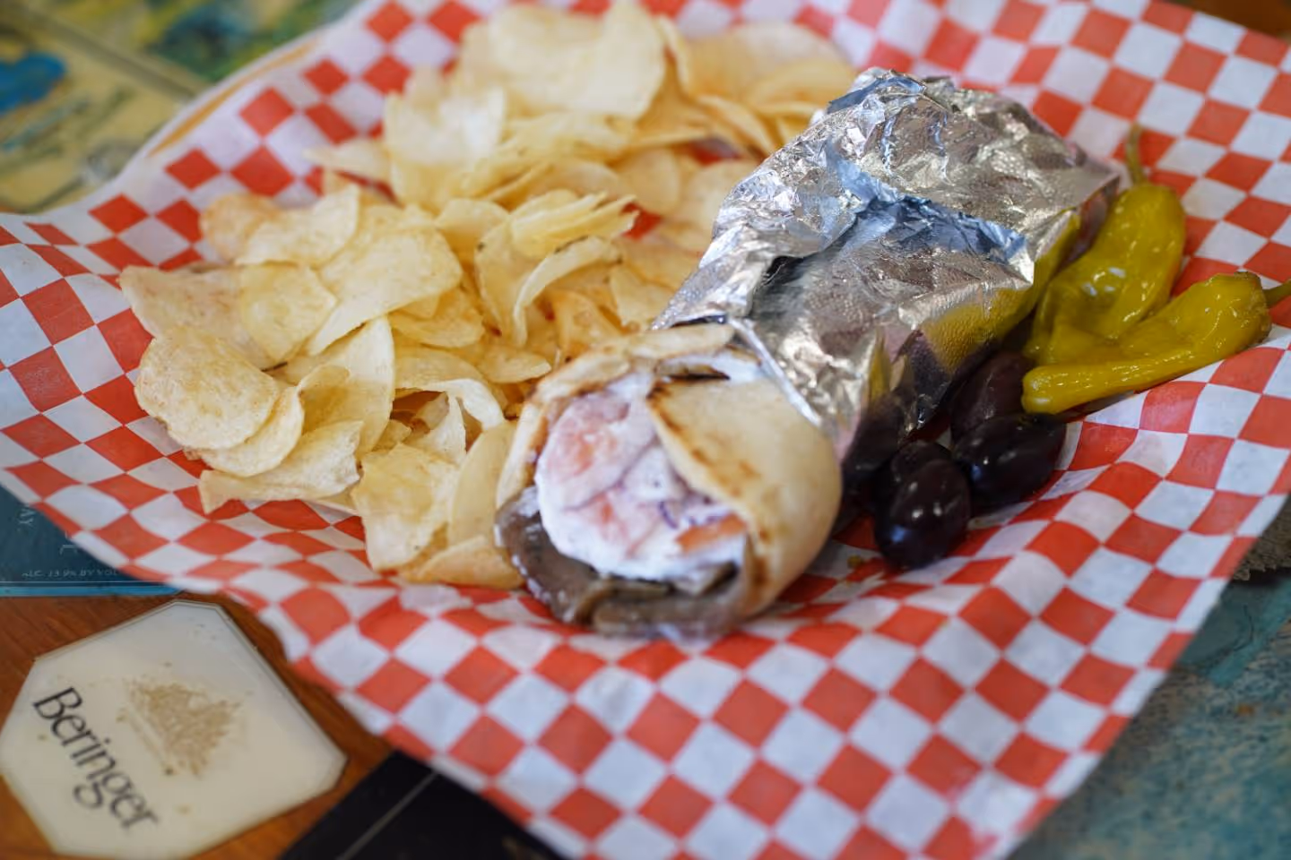 Wrapped gyro with meat and sauce, served with potato chips, olives, and pepperoncini on checkered paper.