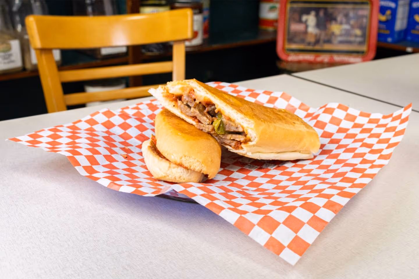 Italian sausage sandwich with meat and peppers on checkered paper on a table with a wooden chair behind.