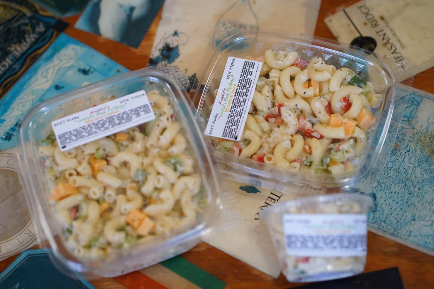 Three containers of macaroni salad with cheese, celery, and peppers on a printed table surface.