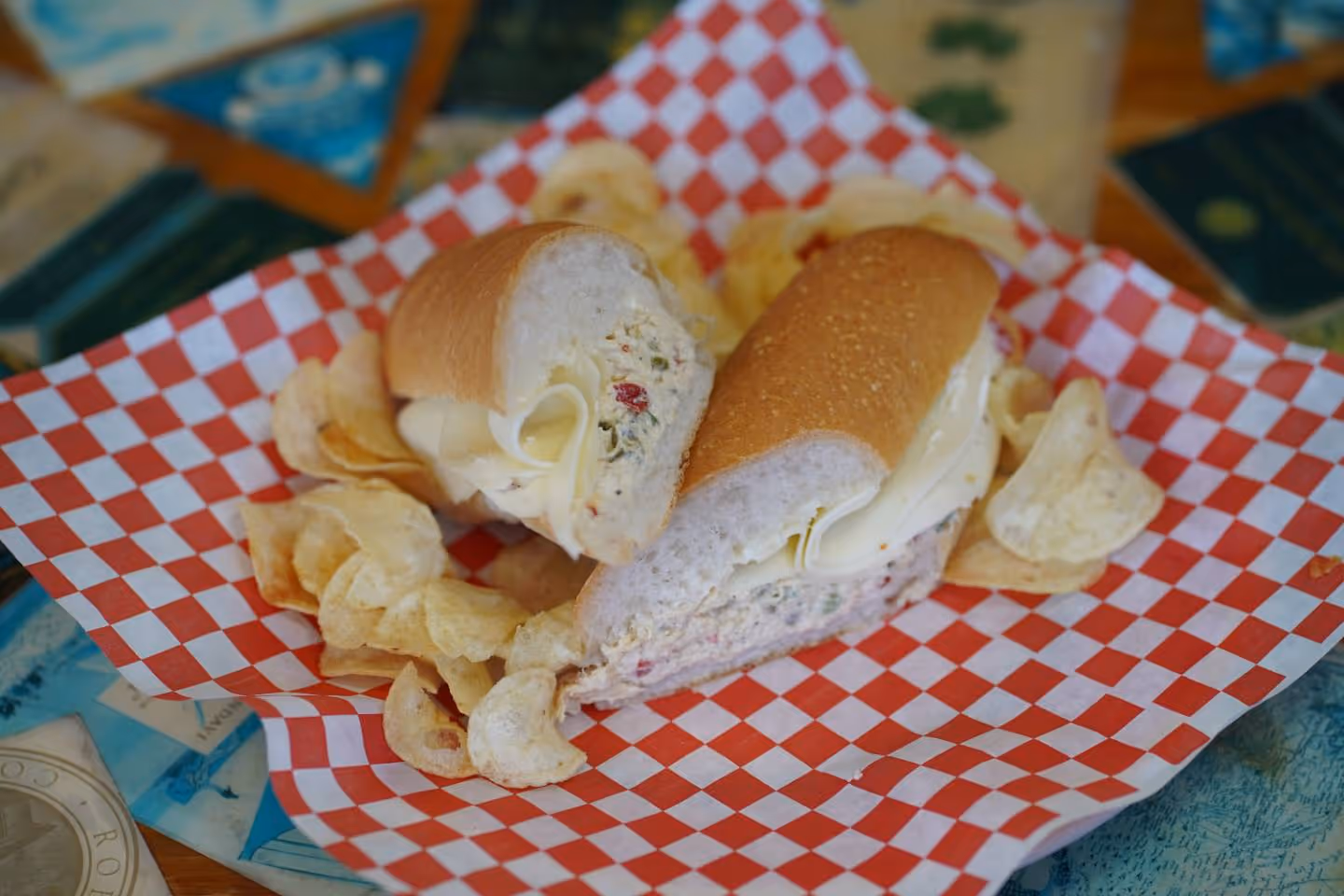 Chicken salad sandwich with slices of cheese on a red and white checkered paper, served with potato chips.
