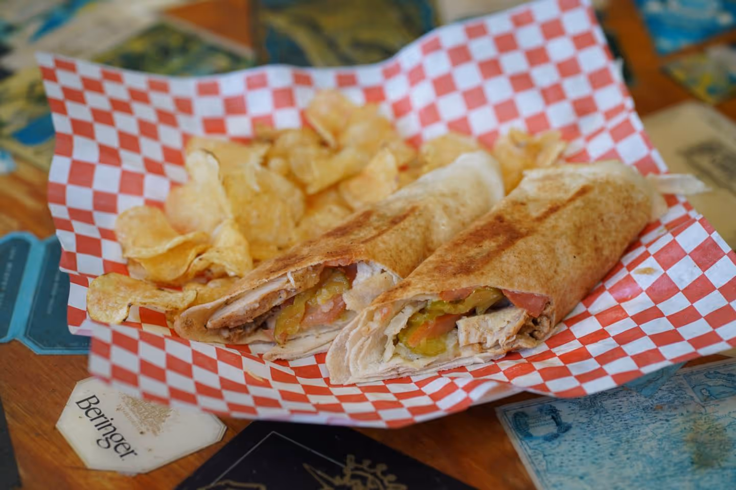 Grilled chicken wrap with pickles and tomatoes served with potato chips on red checkered paper.