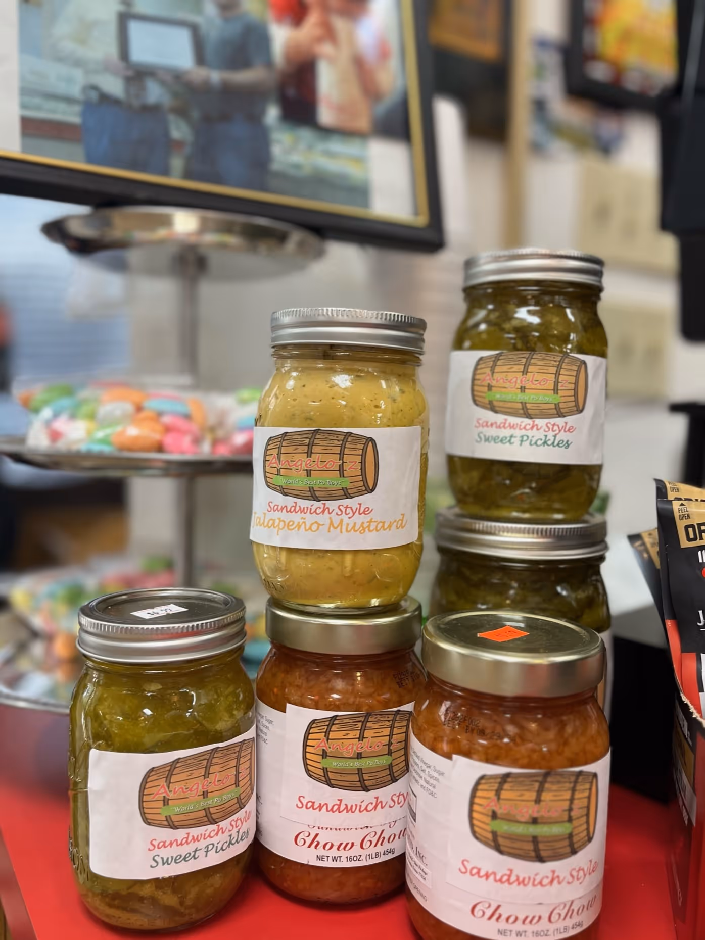 Jars of Angelo Z sandwich-style pickles, jalapeño mustard, and chow chow on a red surface.