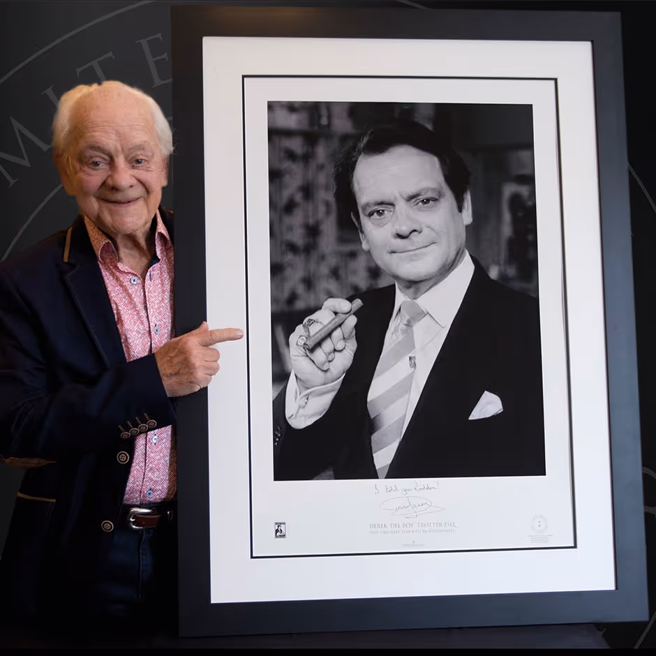 Elderly man pointing at a framed black-and-white portrait photograph while smiling beside it.