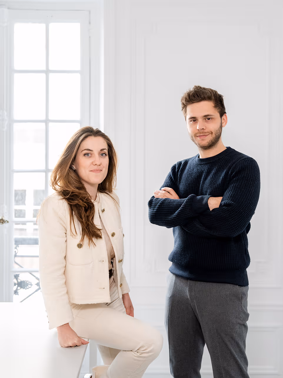 Une femme en tenue claire assise sur un bureau et un homme en pull sombre debout avec les bras croisés dans une pièce lumineuse.