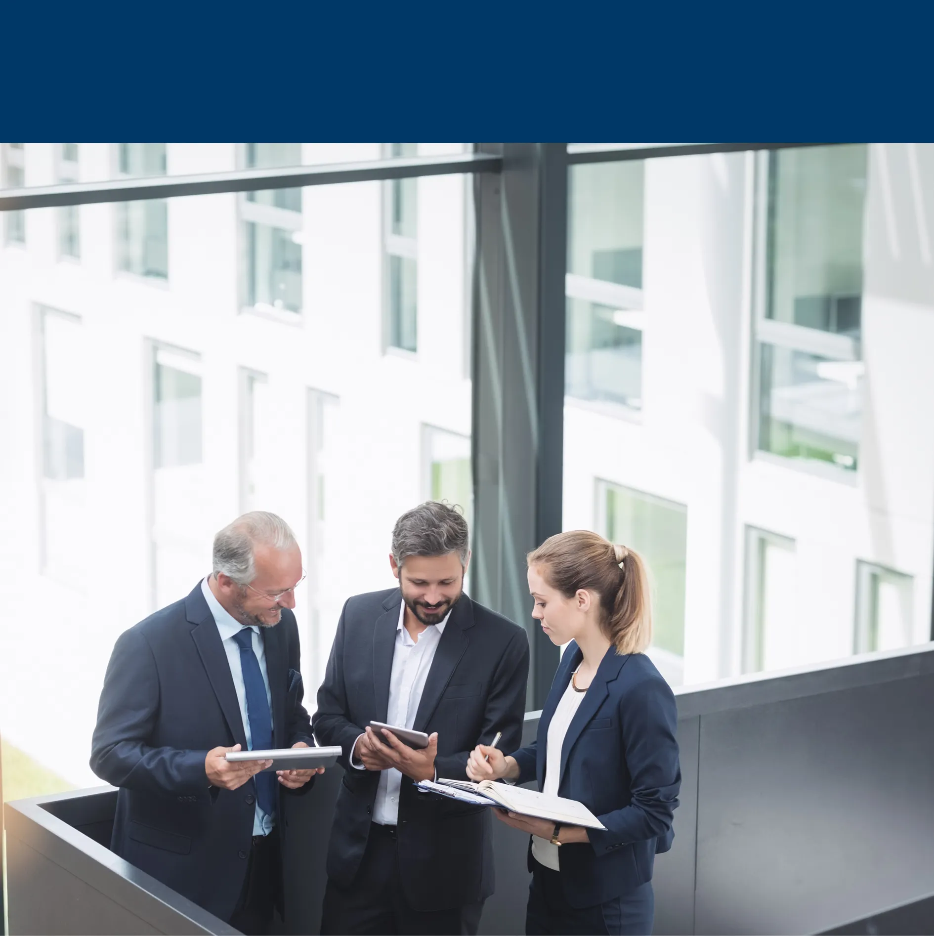 Three business professionals discussing work while holding tablets and documents in an office setting.