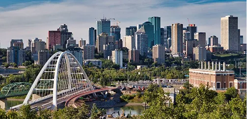 City skyline with modern buildings, a bridge over a river, and green trees in the foreground.