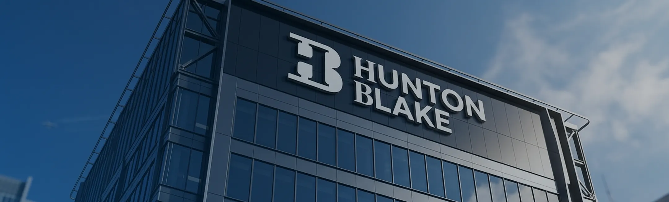 Modern glass office building with large Hunton Blake logo on the upper facade against blue sky.