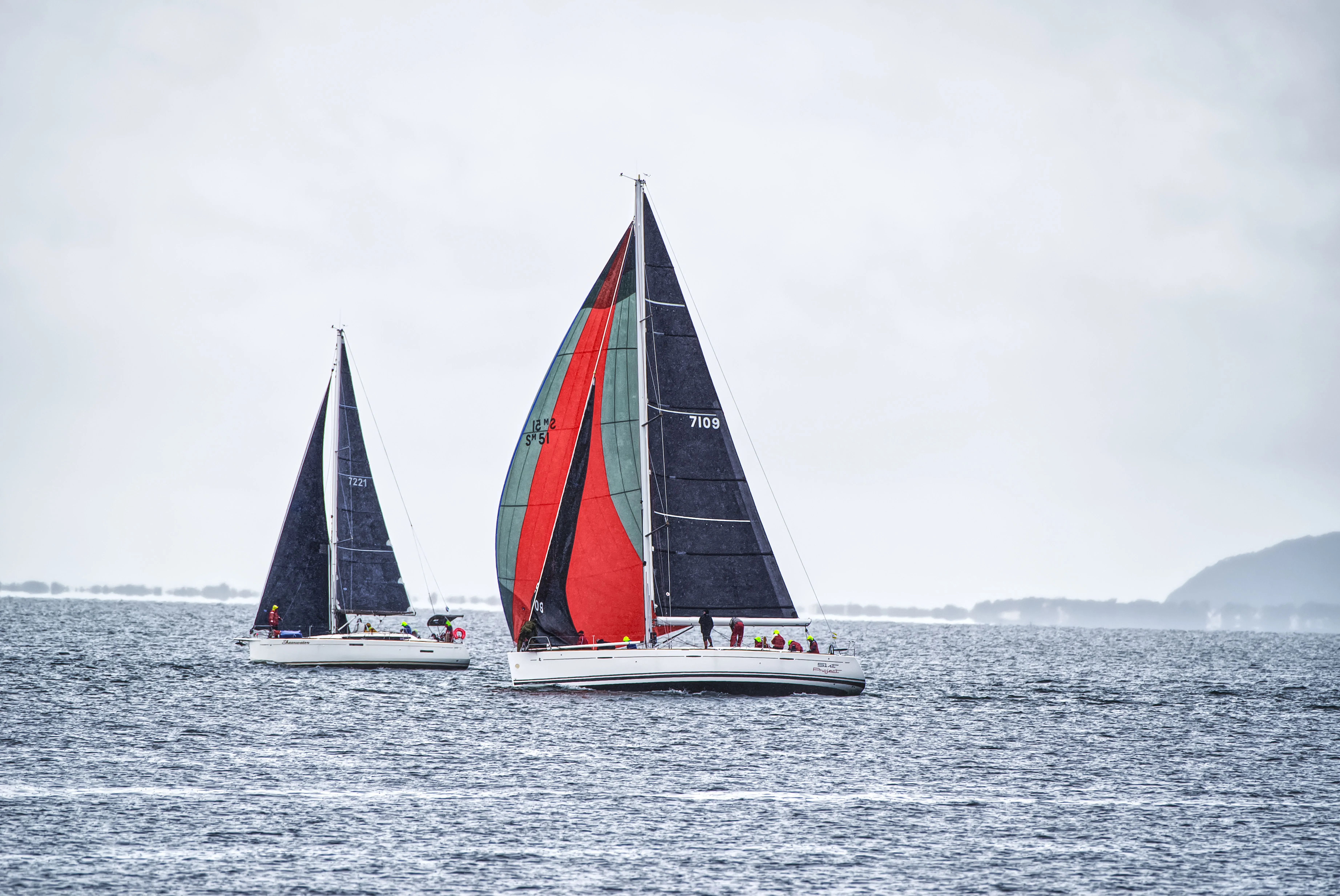 Two sailboats on water with dark sails, one with a red and green spinnaker, under cloudy sky.