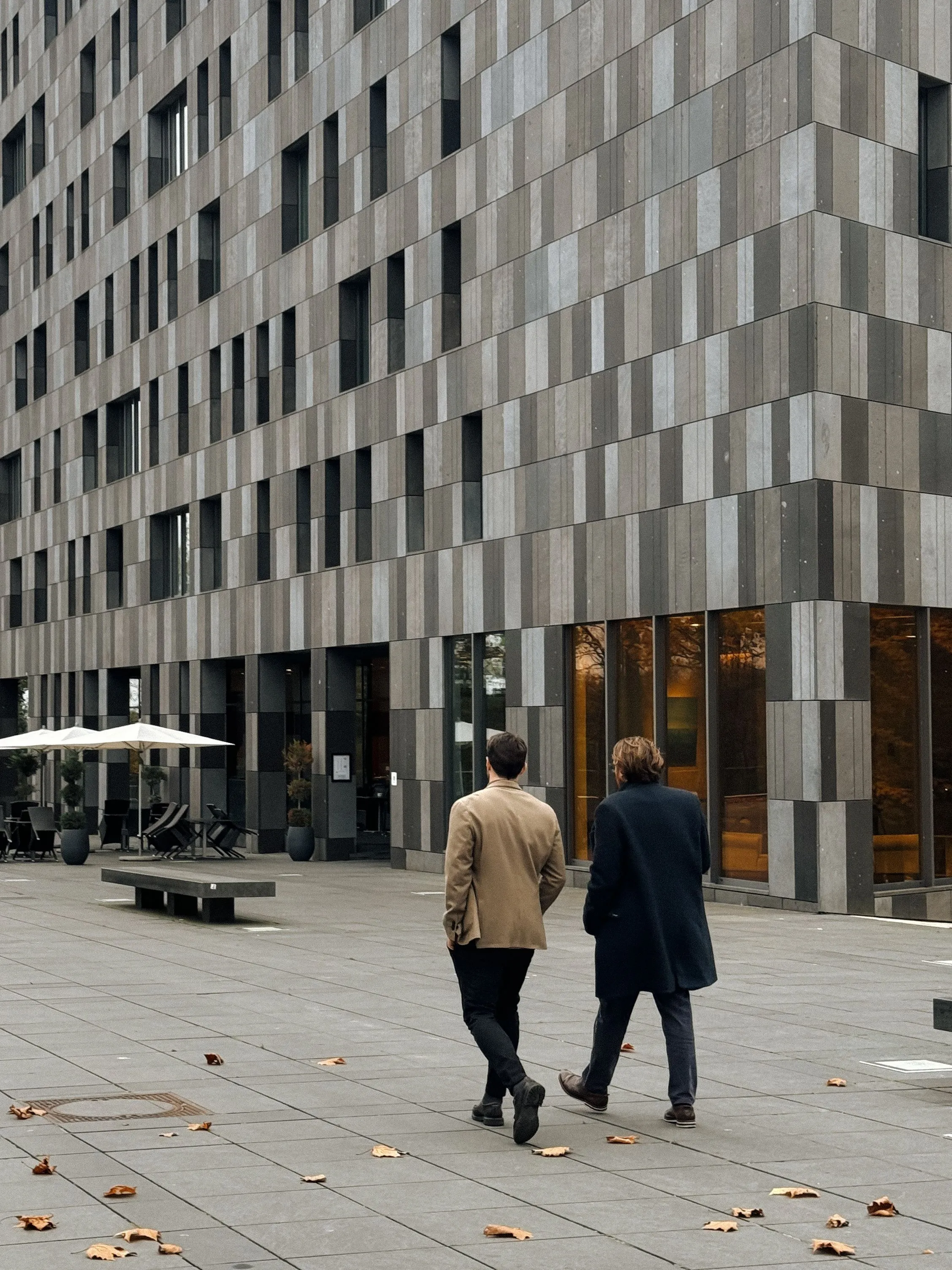 Two men walking near a modern building with gray tiled facade and scattered dry leaves on pavement.