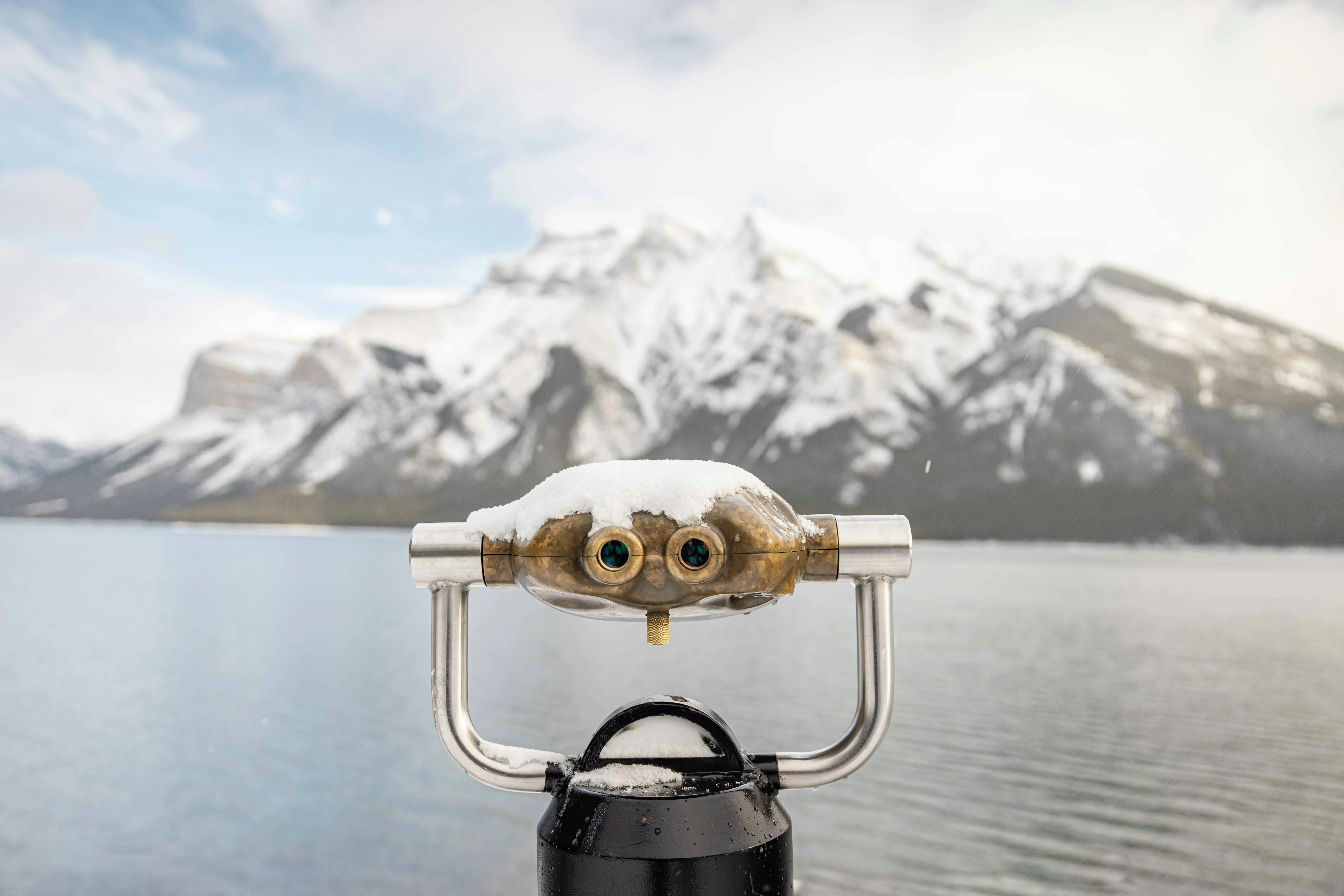 Snow-covered binocular viewer overlooking a lake with snow-capped mountains in the background.