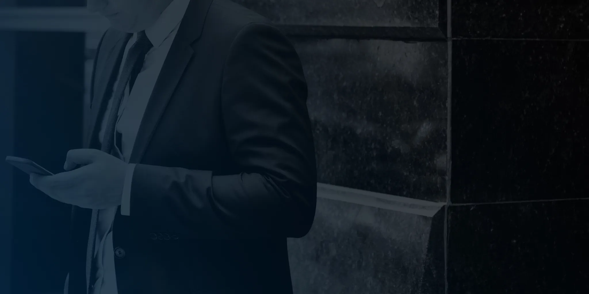 Man in suit using a smartphone while standing against a dark tiled wall.
