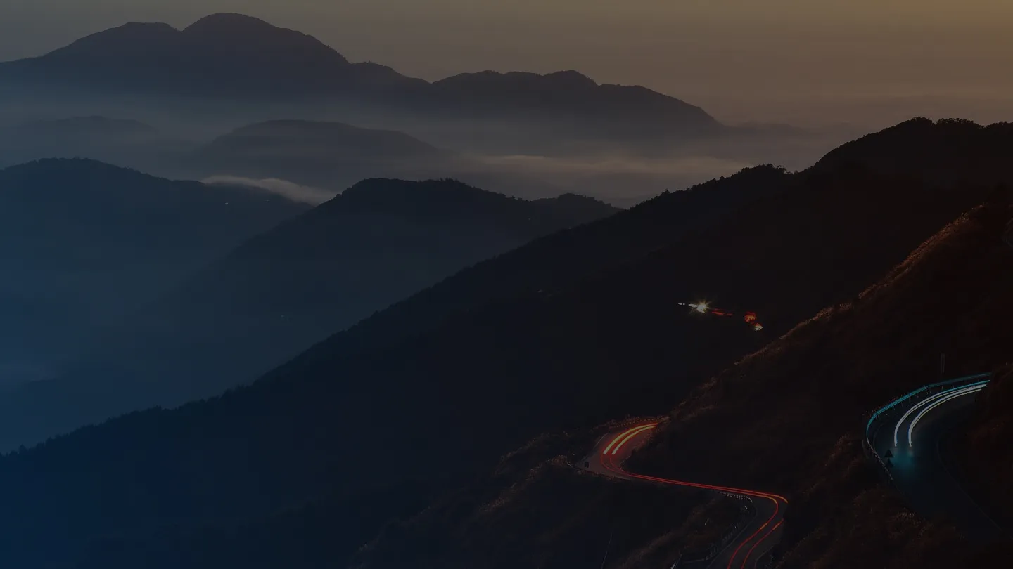 Winding mountain road with light trails of vehicles against misty hills at dusk.