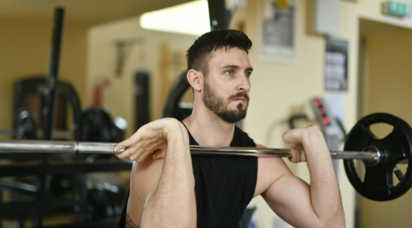 A man in a gym focused on lifting a barbell. He wears a black tank top, showcasing strength and concentration. Fitness equipment is visible in the background.