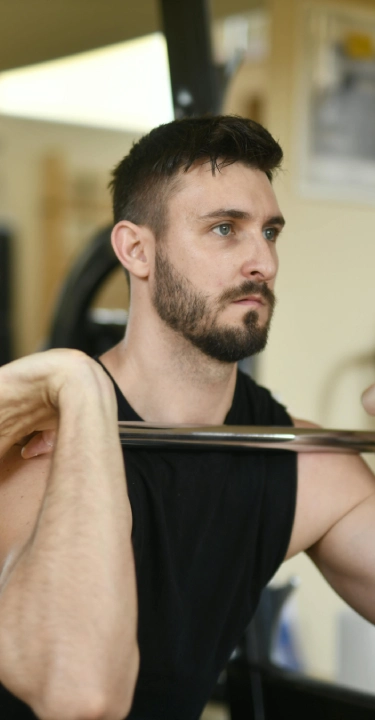 A man in a gym focused on lifting a barbell. He wears a black tank top, showcasing strength and concentration. Fitness equipment is visible in the background.