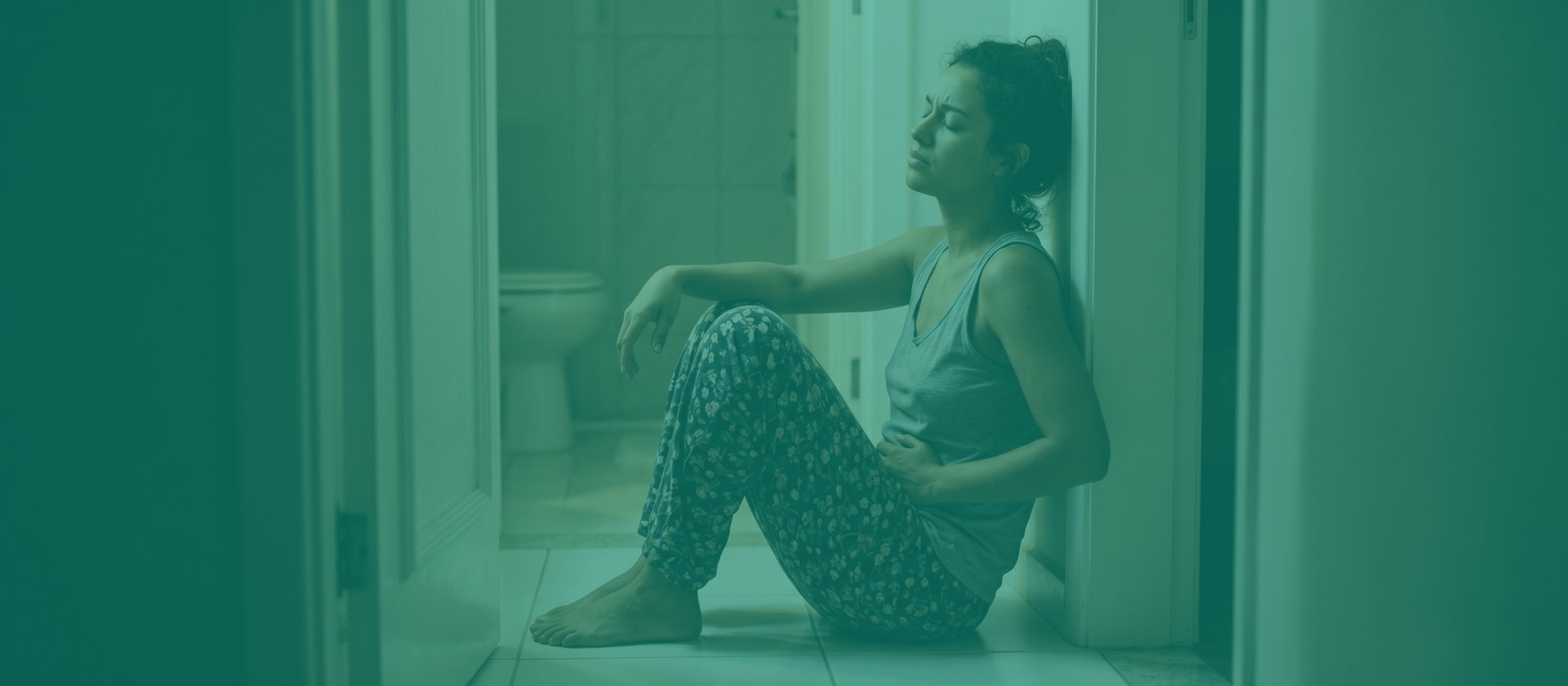 Mulher com virose intestinal sentada no chão do banheiro à noite, encostada no batente da porta, com uma mão pressionando a barriga em cólica