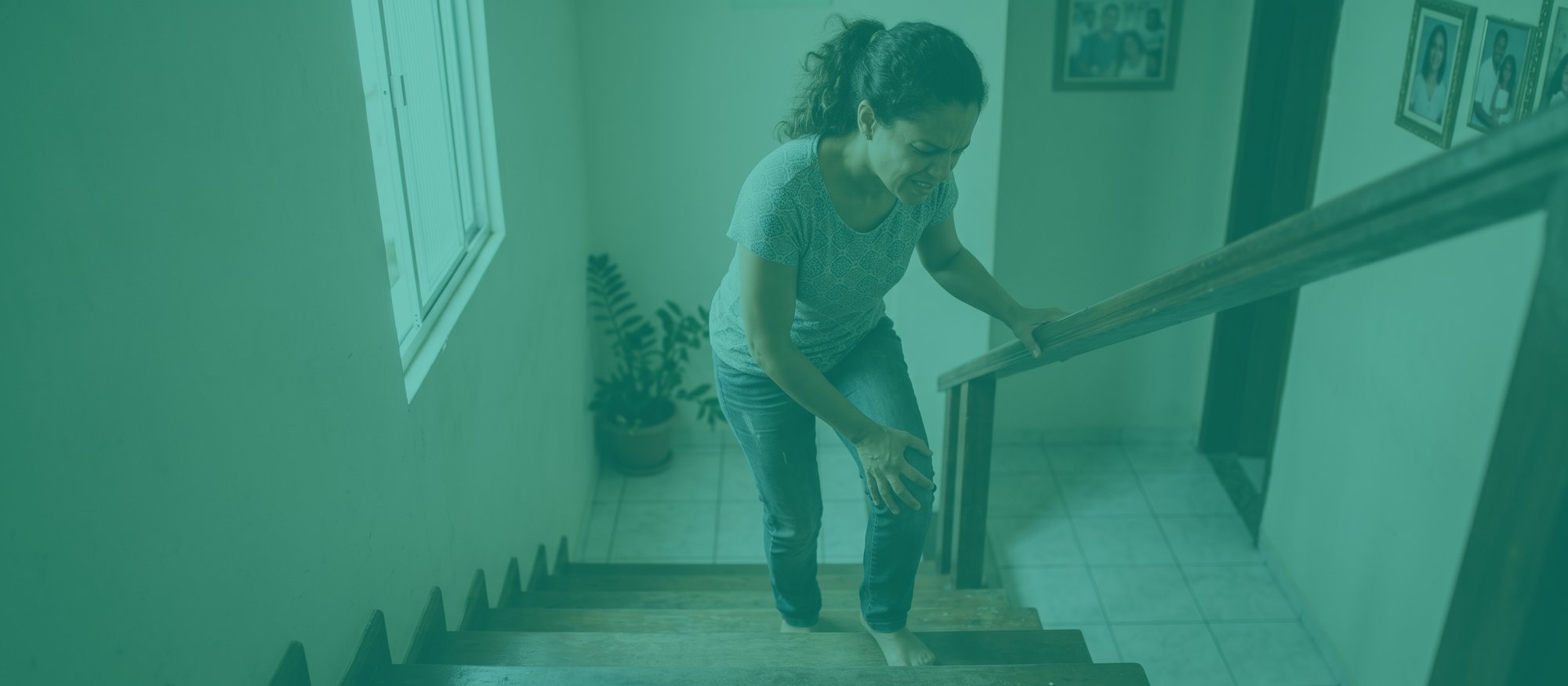 Mulher descendo a escada de casa apoia-se no corrimão e leva a mão ao joelho dolorido, com careta de dor ao pisar