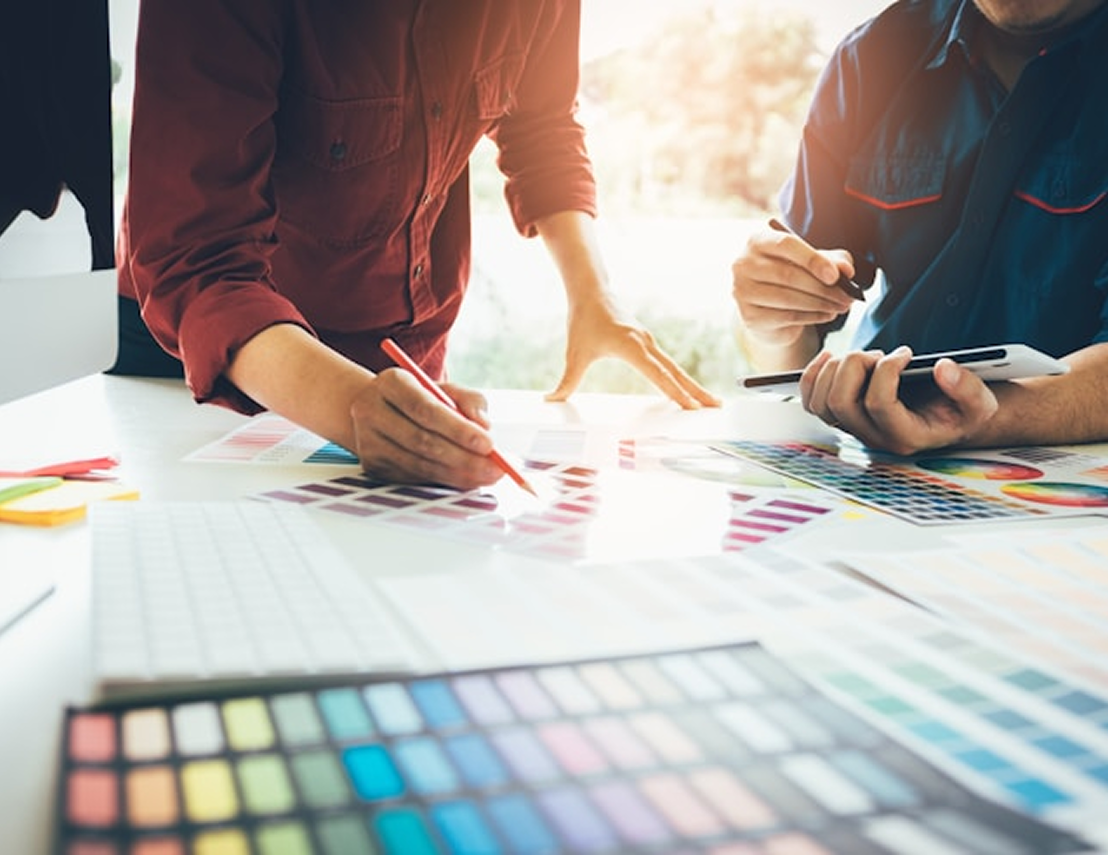 Two people collaborating over a table covered with color swatches and a digital tablet, selecting colors with a pencil and stylus.