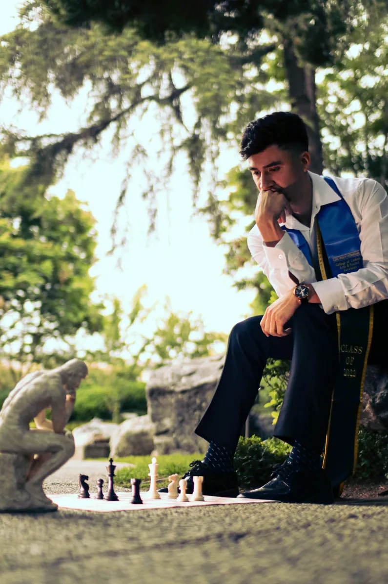 Man in graduation attire sitting outdoors pondering a chessboard next to a thinker statue.