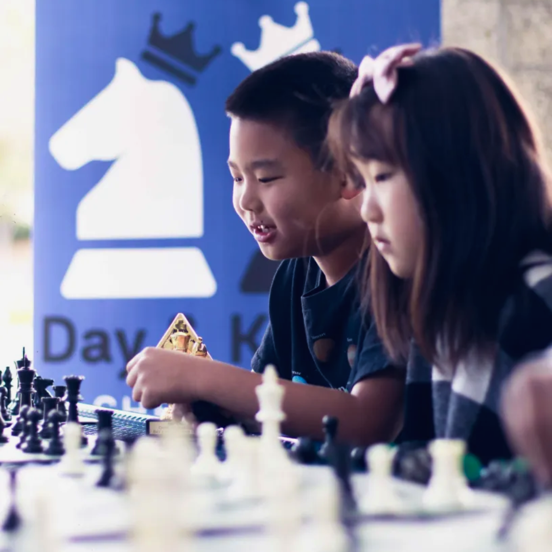 Two children playing chess at a table with a chess-themed blue banner in the background.
