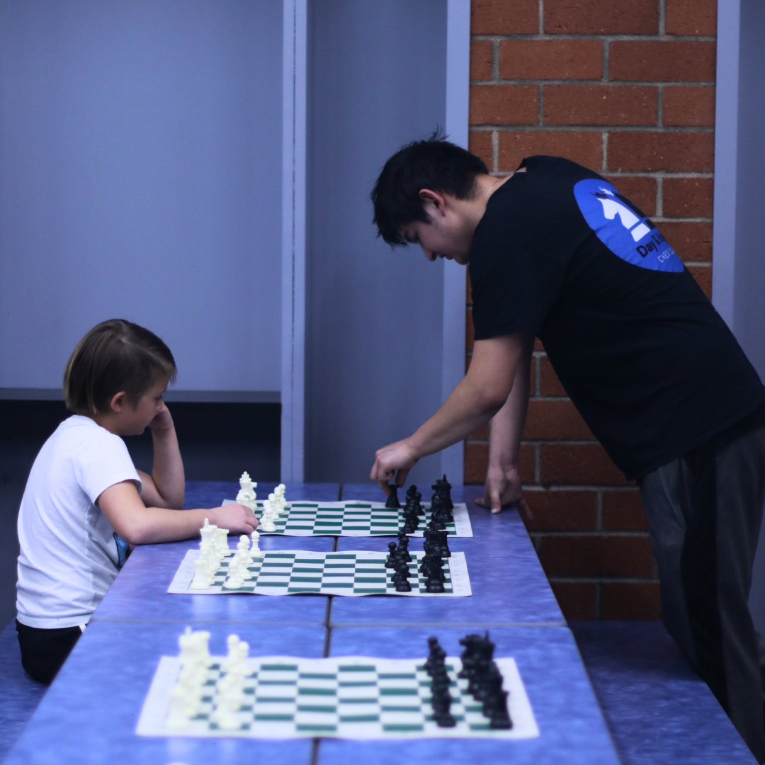 A child and a young man playing chess on multiple boards at a long blue table indoors.