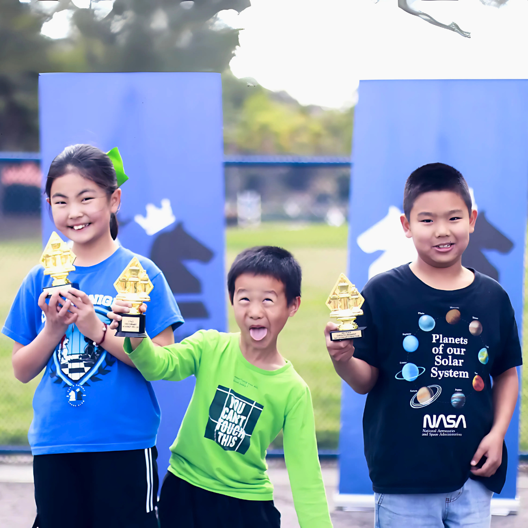 Three children holding trophies, standing outdoors with blue banners featuring chess knight pieces behind them.