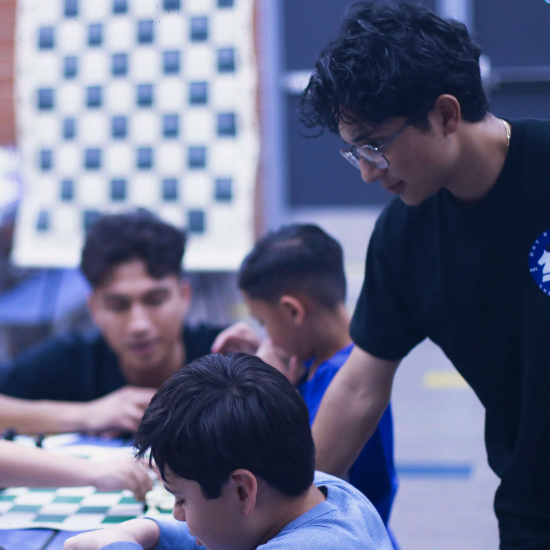 Young man instructing children playing chess with a chessboard visible in the background.