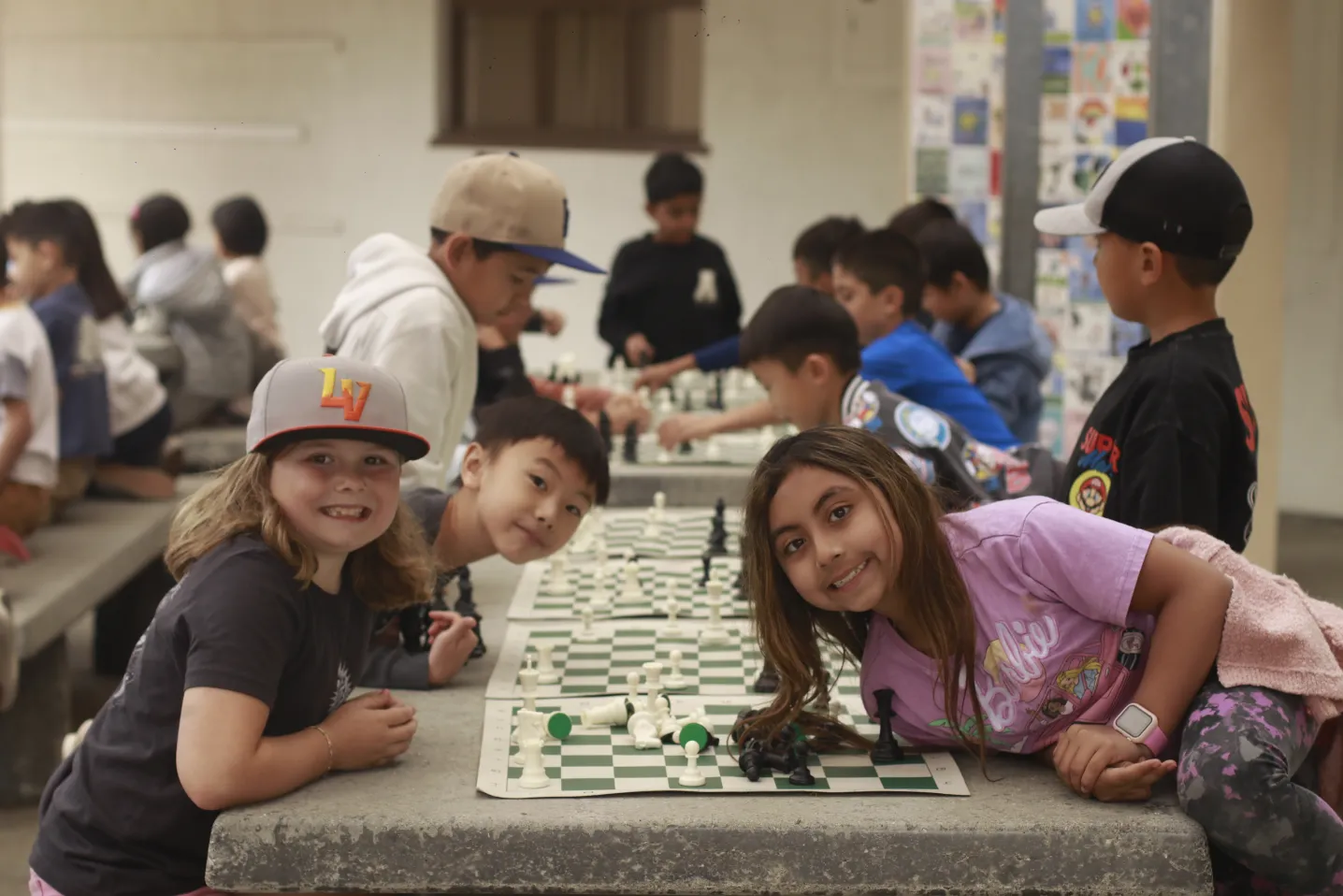 Children playing chess at tables in a group, some smiling at the camera in a casual indoor setting.
