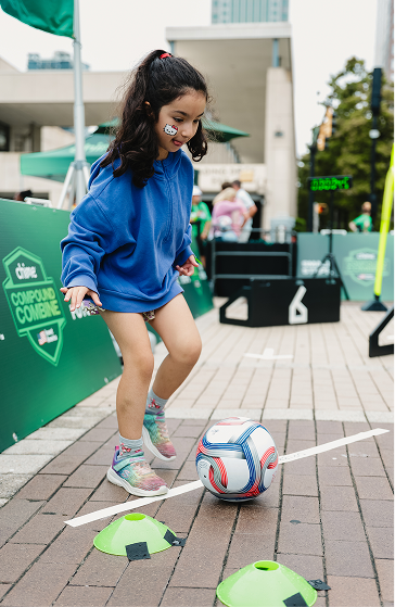 Young girl in blue sweatshirt and shorts dribbling a soccer ball between green cones on a brick pavement during an outdoor event.
