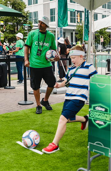 A young boy in a striped shirt and red shoes is kicking a soccer ball on artificial grass while a man in a green shirt and bucket hat watches nearby holding two soccer balls.