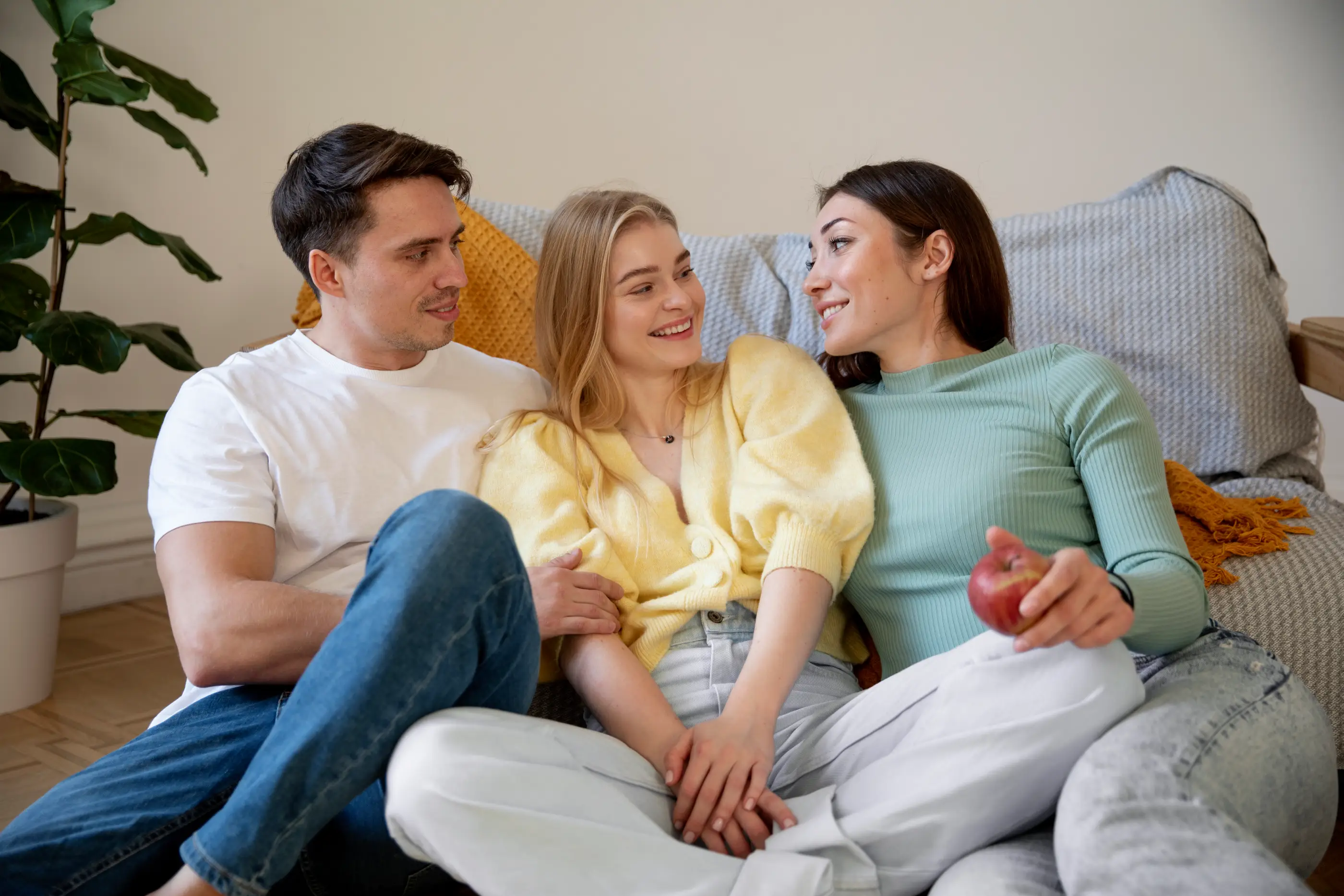 A man and two women sitting closely on a sofa, smiling and looking at each other warmly.