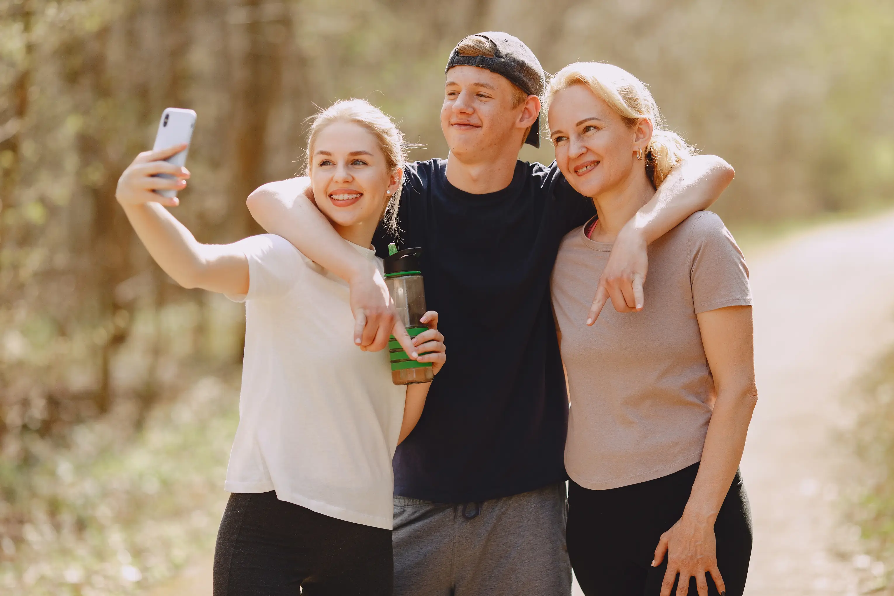 Young man with cap embraces two women; one takes a selfie while holding a water bottle outdoors.