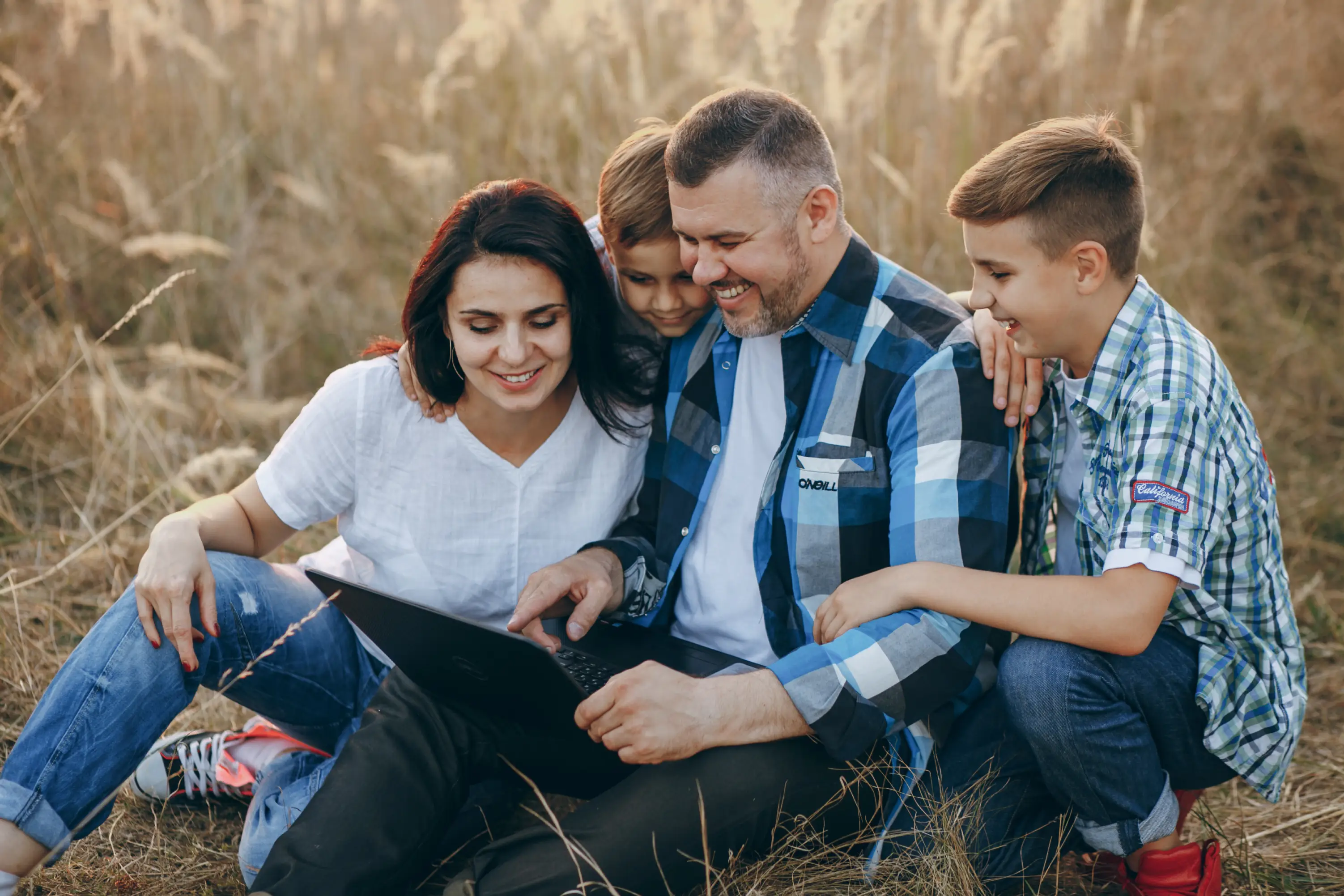 Smiling family of four sitting in a field looking at a laptop together in casual clothes.